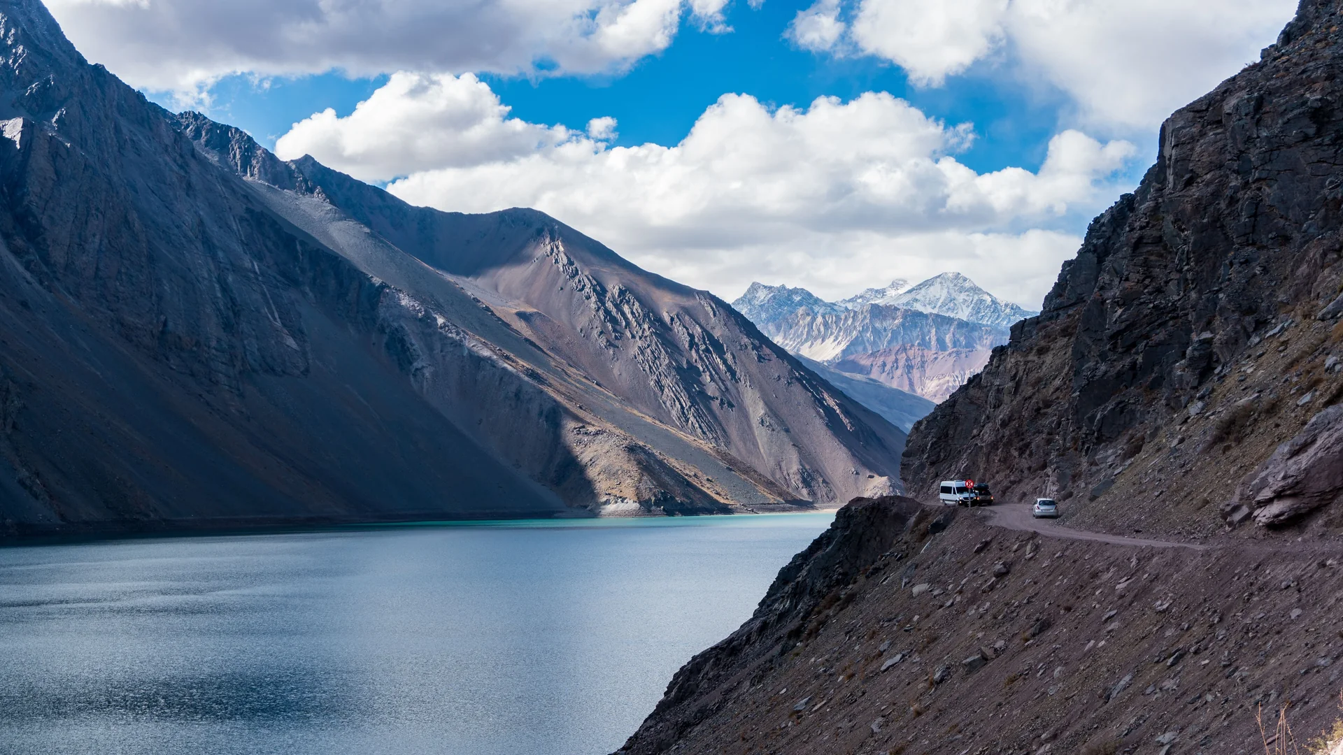 En varevogn og en bil på en smal grusvej med hårnålesving over det turkisblå Embalse El Yeso-reservoir, omkranset af nøgne andinske tinder
