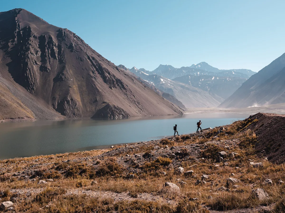 To vandrere, der går langs bredden af den turkisblå Embalse El Yeso-reservoir under nøgne andinske tinder
